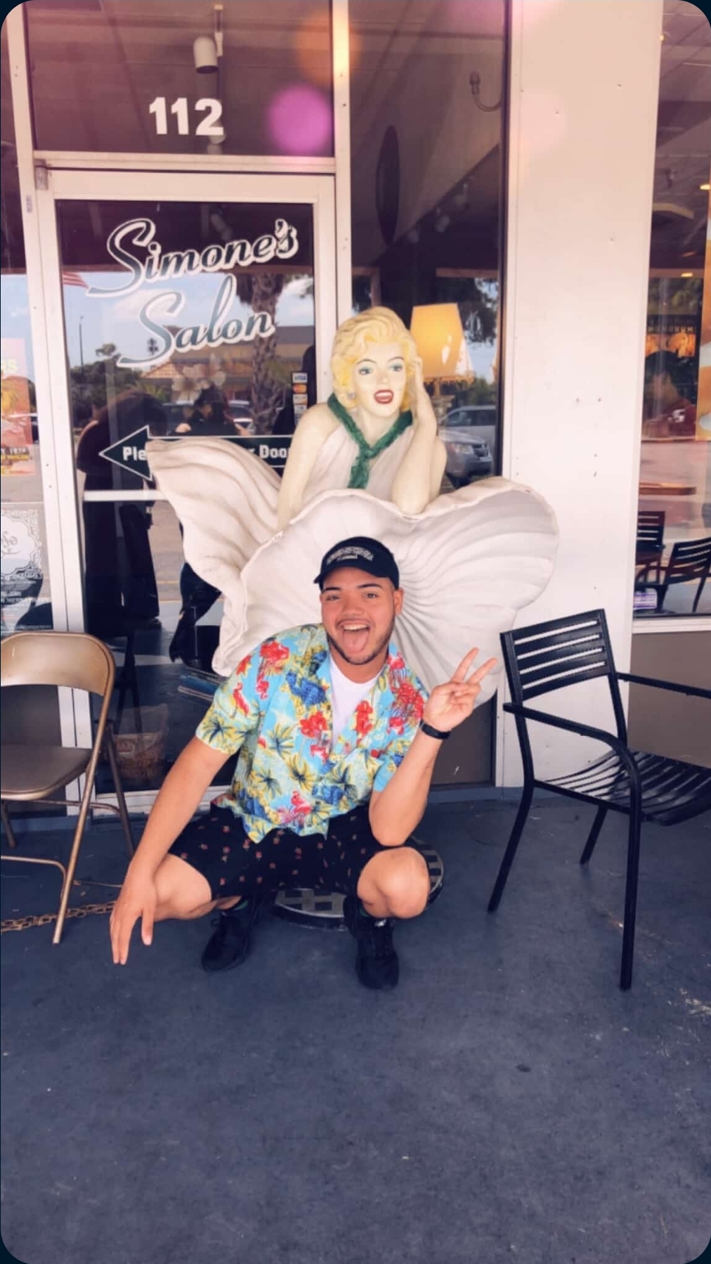 Author Manuel Cross crouching and making a peace sign in front of a storefront, with a Marilyn Monroe statue behind him.